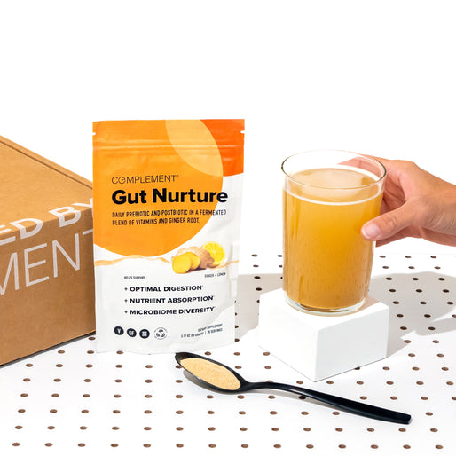A hand holds a glass of orange beverage next to a packet labeled Gut Nurture and a black spoon filled with powder, all placed on a white, dotted surface with a cardboard box in the background.