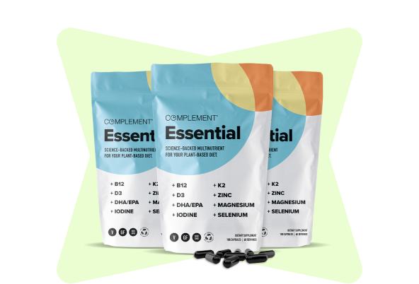 Three bags of Complement Essential multivitamins are displayed in a row. The packaging highlights nutrients like B12, D3, DHA/EPA, K2, zinc, magnesium, iodine, and selenium. Black capsules are shown in front of the bags. The background is light green.