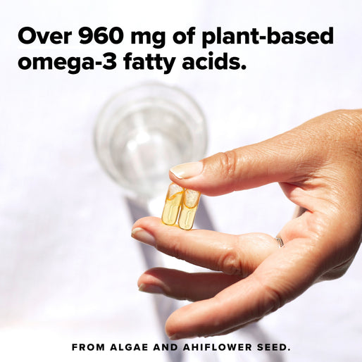 A hand holds two clear capsules above a white surface with a glass of water nearby. Text reads: Over 960 mg of plant-based omega-3 fatty acids. From algae and ahiflower seed.