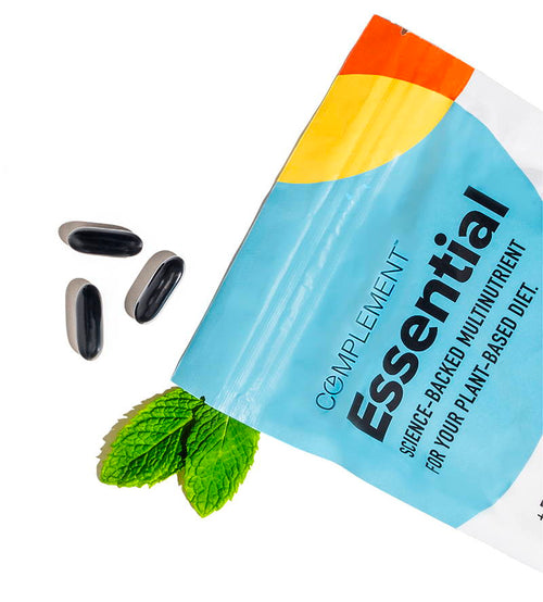A blue and orange resealable pouch labeled Complement Essential with text highlighting plant-based nutrition. Four capsules lie beside the bag, next to sprigs of fresh mint leaves.