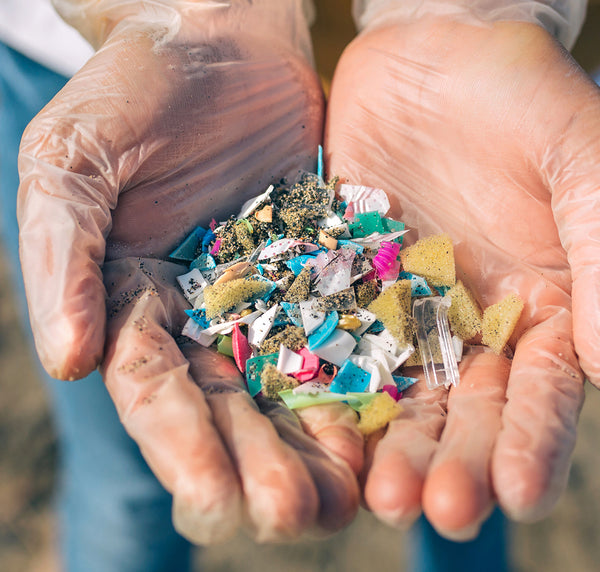 A person wearing clear gloves holds a handful of colorful plastic fragments and sand, highlighting pollution and environmental concerns.
