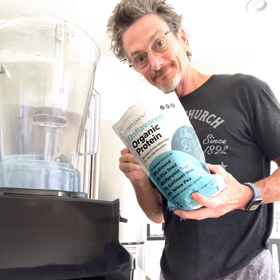A smiling man wearing glasses and a dark t-shirt holds a bag of unflavored organic protein powder next to a blender in a bright kitchen.