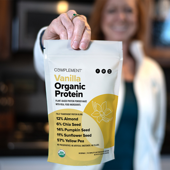 A person holds a bag of Complement Vanilla Organic Protein powder, featuring its plant-based ingredients and nutrition facts on the front, with a blurred background.