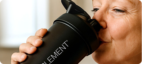 A person smiles while drinking from a black shaker bottle labeled SUPPLEMENT. The background is softly blurred.