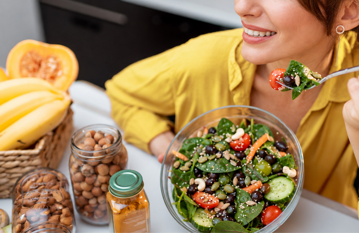 A person in a yellow shirt holding a forkful of salad from a bowl, with jars of nuts and a basket of produce nearby.