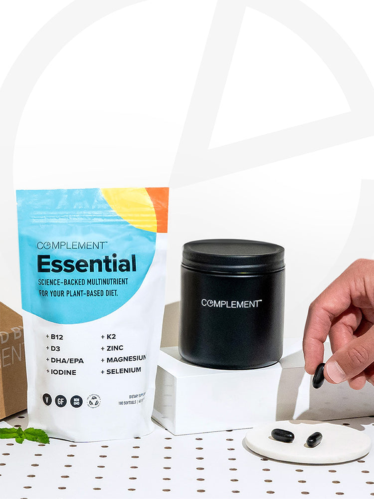 Hand holding Complement Essential vegan multivitamin next to the pouch, matte black glass jar for storage, and shipping box. 