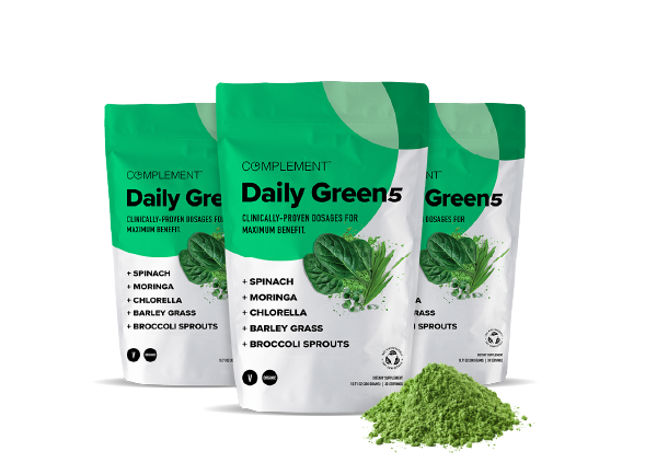 Three green and white bags of Complement Daily Green 5 powder are displayed, featuring spinach, moringa, chlorella, barley grass, and broccoli sprouts. A pile of green powder is in front of the bags.